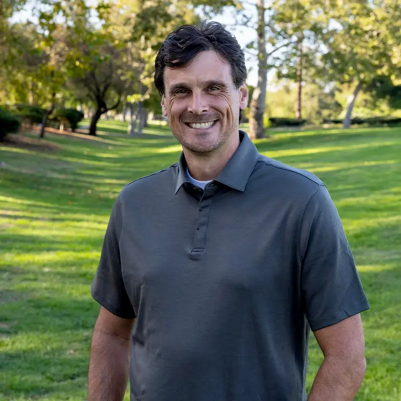 Chris Kluwe smiling in front of a green park and trees.