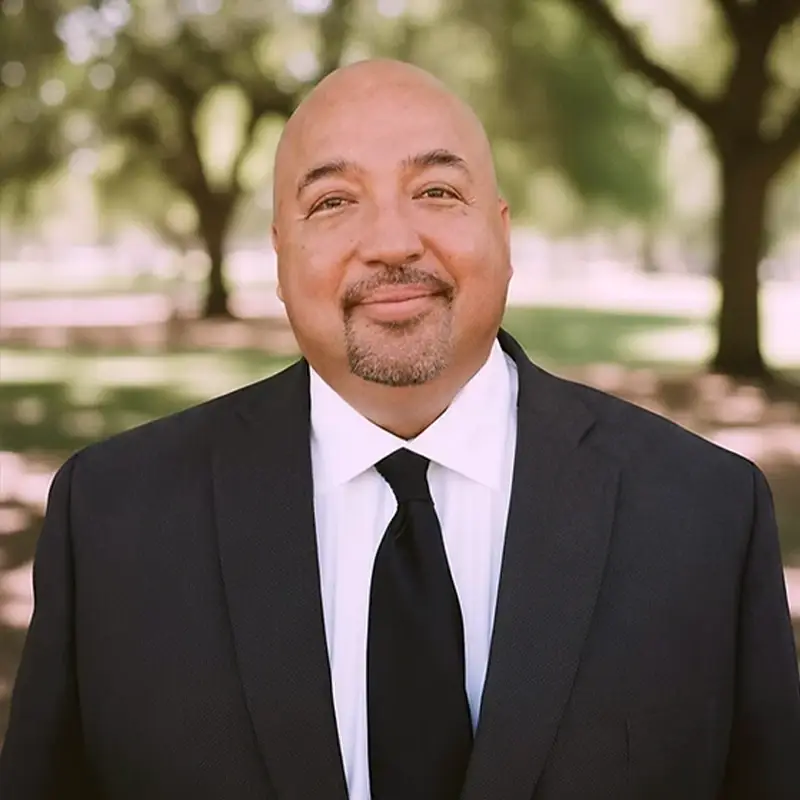 Andy Godinez, wearing a suit, smiling in front of trees and greenery.