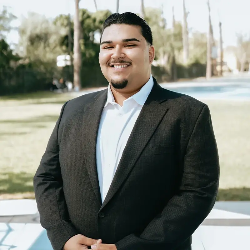 Hector Bustos, smiling wearing a blazer in front of a sunny park.