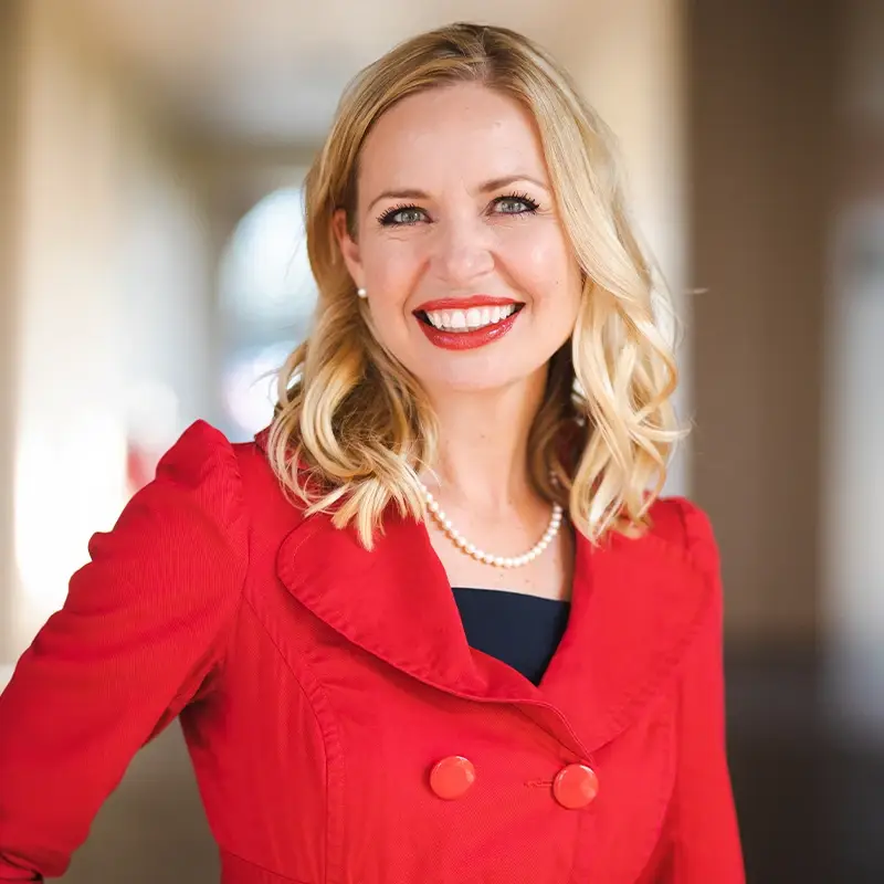 Ruthi Hanchett, smiling in front of a corridor wearing a red coat.